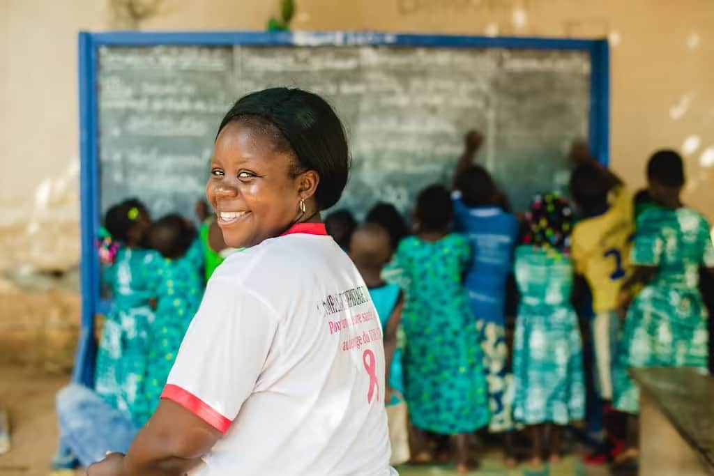 Une femme togolaise est assise dans une salle de classe où des enfants travaillent sur un tableau à l'arrière-plan.