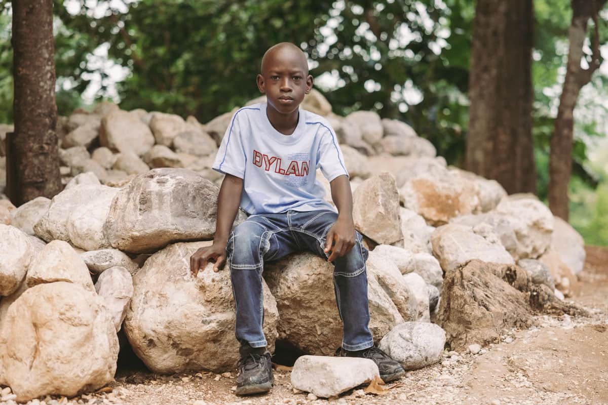 Haiti: un bambino in maglietta e jeans siede su un mucchio di pietre.