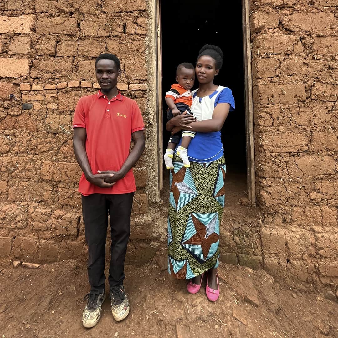 une participante au programme de survie se tient devant sa maisonnette en argile avec son mari et son bébé.