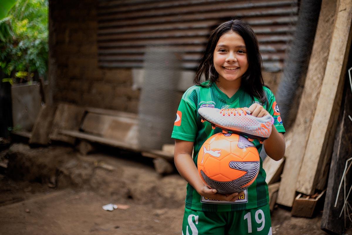 Karla, una bambina sostenuta da Compassion, fuori casa con un pallone da calcio arancione e un paio di scarpe da calcio.